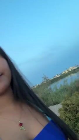 A woman with long dark hair, wearing a blue and turquoise tie-dye dress and a necklace with a red rose pendant, is walking outdoors. She is looking at the camera and sticking out her tongue. The background features a body of water, vegetation, and a cloudy sky. The ground appears to be gravelly. The video captures the woman in motion, with some blurring indicating movement.