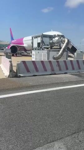 The video shows an airport apron on a sunny day. Initially, a Wizz Air airplane is visible with a mobile air conditioning unit attached. The camera pans right to show a Ryanair aircraft being serviced. Baggage carts and a small service vehicle are parked near the plane.  The sky is blue with some white clouds.