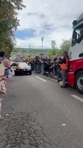 The video shows a black Bugatti Chiron with orange racing stripes moving slowly down a cobblestone street lined with crowds behind barriers. A large red and white truck is parked on the right side of the road. A man wearing an orange vest stands next to the truck. People in the crowd are taking photos and videos of the car. A man and a woman are visible inside the car. The weather is overcast. Buildings and trees are visible in the background.
