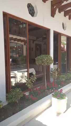 The video pans across an outdoor courtyard with an archway overhead. The courtyard has a light-colored paved floor. To the left, there is a building with large windows framed in dark wood, a flower bed with red and pink flowers, and a small round table with a chair. The building has an overhang supported by wooden beams. Further into the courtyard, there are people walking and a set of stairs leading up to a higher level with more buildings and decorative wooden railings. The sky is blue with some white clouds.