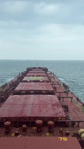The video shows a perspective from the deck of a large cargo ship sailing on the ocean. The ship is moving forward, indicated by the wake visible in the water on either side. The deck is covered with large, red, rectangular hatches that conceal the cargo holds. There are yellow markings on the deck, including the numbers "6S" and "7S." In the distance, the horizon is visible, with a cloudy sky overhead and a few other ships in the far distance. The sea appears to be somewhat choppy. The overall impression is one of a vast, open ocean and the immense scale of maritime transport.