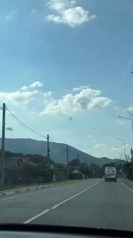 A slightly shaky, first-person perspective video captures a view from inside a vehicle traveling on a paved road. The road has white lane markings. To the left of the road are houses with roofs, trees, and utility poles with wires. In the distance, rolling hills or mountains are visible under a bright blue sky with scattered white clouds. A white van is seen ahead on the road, moving in the same direction as the camera. Traffic lights and streetlights are also present along the road. There are some dark specks on the camera lens or windshield. The time stamp "00:00" is displayed at the beginning of the video.