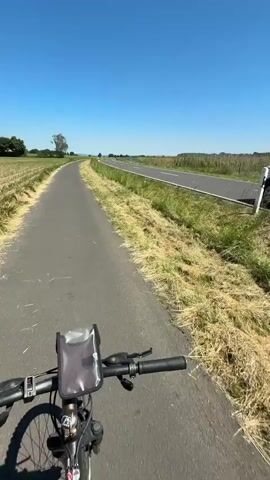 The video shows a young woman riding a bike on a paved path next to a field of crops under a clear blue sky. A road with cars driving on it runs parallel to the bike path, separated by a grassy median and fence. As the video begins, the camera faces forward along the path.  Later, the woman is visible wearing sunglasses, a black top, and black shorts. She turns the camera towards herself to show her smiling face and give a peace sign to the camera.