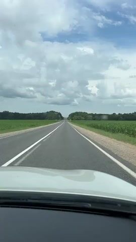 This is a first-person view from a vehicle driving down a straight, two-lane asphalt road. The road has a white solid line on the right edge and a white broken line separating the two lanes. On either side of the road are agricultural fields with green crops, possibly corn. Beyond the fields, there are rows of trees forming a distant treeline. The sky is partly cloudy with white cumulus clouds against a blue sky. The front of the vehicle, including the dashboard and hood, is visible in the foreground.