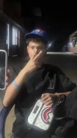 A young man in a baseball cap, dark t-shirt, and black pants sits outdoors at night, facing the camera. He holds up a hand with three fingers showing, then points down. The camera briefly zooms in on his face. The environment is dark with a window and streetlights visible in the background. The man is also holding a small, patterned bag.