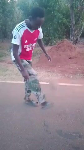 A young man stands on a paved road wearing a red and white sports jersey with the words "Emirates Fly Better" on it, along with camouflage pants and sandals. He is juggling a soccer ball with his feet, counting aloud to 11. A pile of dirt and vegetation can be seen on the side of the road.