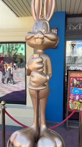 A shiny, gold-colored statue of Bugs Bunny stands outside a building. The statue has a carrot held in front of it. Red rope is used as a barrier in front of the statue. People walk by the building in the background. To the statue’s right, an advertisement for ice cream stands on the sidewalk.