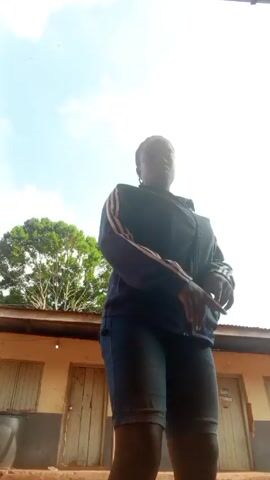 A medium shot from a low angle shows a person standing in front of a low, one-story building with three visible closed doors. The building is a pale orange color with a dark roof. A large, leafy green tree is partially visible behind the building and to the left. The person is wearing a dark-colored zip-up jacket with white stripes on the sleeves and denim capri pants. The person is facing the camera and gesturing with their hands. The sky is light blue with some clouds. A motorcycle passes in the background.
