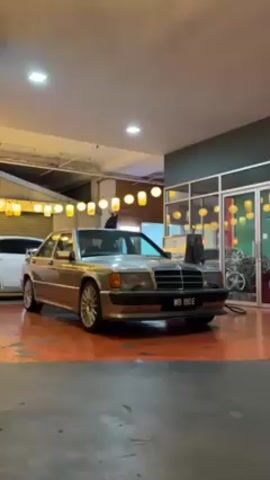 Here is a description of the video:

The video shows a light tan Mercedes-Benz 190E parked inside a garage or workshop. The car is positioned at an angle, facing the camera, and its headlights and grill are clearly visible.  The front license plate reads "WB 190E". Behind the car, along the left wall, is a white minivan. A string of decorative lights hangs from the ceiling above the car. In the background, a person is briefly visible in the doorway of a glass-enclosed room or office. Inside that room, various car parts and wheels can be seen. The floor of the garage is orange.