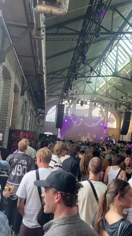 The video shows a crowd of people inside a large industrial-style building. The ceiling is high with exposed beams and skylights. There is a stage at the far end of the building with a band performing, illuminated by purple and pink lights. The crowd is dense and appears to be attending a concert or music festival. Many people are standing, some are holding drinks, and some are talking to each other. The atmosphere seems lively and energetic. The camera is positioned at ground level, looking towards the stage, and the focus is on the crowd in the foreground.