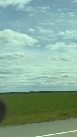 The video shows a view from inside a car traveling on a road. The landscape outside the car includes a large green field and a sky filled with clouds. The camera focuses on the window and then moves to show the seatbelt, the dashboard, the steering wheel, and the side mirror of the car. Another car is visible in the distance on the road ahead.