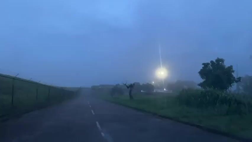 The video displays a static, wide shot of a fog-enshrouded outdoor scene during low light conditions. An asphalt road with a white dashed center line extends from the foreground into the hazy distance, flanked by green grass and vegetation. A barbed wire fence runs along the left side of the road. To the right of the road, a bare, gnarled tree trunk stands. A prominent street light on the right side brightly illuminates a localized area, piercing through the fog. In the far background on the right, several tall, indistinct structures, likely communication towers, are partially obscured by the dense fog. One of these towers repeatedly flashes a bright orange light, visible at approximately 0:01 and 0:02. No people or animals are visible, and no discernible interaction occurs beyond the flashing light.