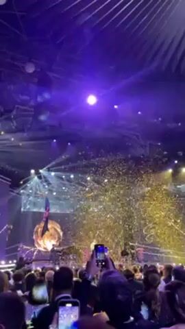 The video captures a performance on a stage with an audience in the foreground. The stage features a large, spherical structure on the left and a video screen in the background. Gold confetti rains down from above, creating a sparkling effect throughout the scene. A person hangs from a rope attached to the ceiling, above the large sphere. The audience is packed closely together, with many people holding up their smartphones to record the event. The lighting is dramatic, with purple spotlights illuminating the scene.