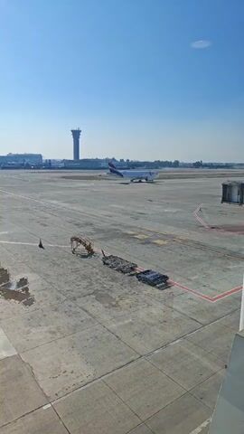 The media file captures an outdoor scene at an airport, likely during daytime due to the clear blue sky and bright sunlight. The primary subjects are an airplane on the tarmac, a control tower in the distance, and various airport equipment such as baggage carts and a service vehicle.

The airplane, appearing to be a commercial jet with a white fuselage and blue and red accents on its tail, is positioned on a runway or taxiway. In the background, a tall, distinctive control tower stands prominently against the sky, indicating the presence of an active airport. Scattered on the tarmac are several pieces of ground support equipment, including what appears to be a baggage conveyor belt system and a utility vehicle with an orange boom. The ground is paved with concrete, marked with yellow and red lines, typical of airport operational areas.

The setting is an airport tarmac on a clear, sunny day. The vast expanse of concrete and the presence of an airplane and control tower strongly suggest an airport environment. The bright sunlight and clear sky imply fair weather. There is no visible text in the media file. The overall mood is one of routine airport operations, with the airplane poised for movement and the infrastructure in place to support air travel.