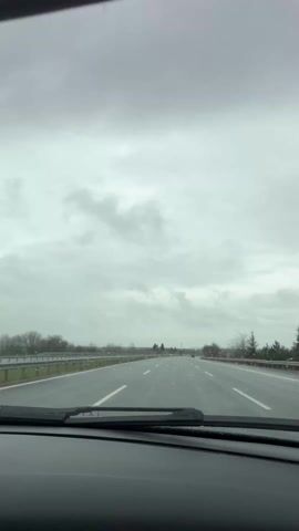 The video shows the view from inside a car driving on a highway. The sky is overcast and gray, and there is light rain. The windshield wipers are on, wiping away the raindrops. The road ahead is straight, with three lanes visible. There are trees and fields on either side of the road. A few other cars can be seen in the distance.