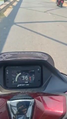 A first-person perspective video shows a person riding a scooter across a bridge. The scooter's digital dashboard displays a speed of 36 km/h. The bridge has metal railings and a river runs below. Another person is seen walking further ahead on the bridge, and a white car is also visible. The sun is shining, creating shadows on the bridge.