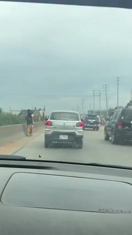 A video captures a street scene from inside a vehicle, showing traffic on a cloudy day. In the foreground, the dashboard of the car is visible. On the left side of the road, a concrete barrier lines the side, with several individuals standing on top of it or near it. One person, possibly a woman, is holding a child. Further down the barrier, two younger individuals, a female in yellow pants and a male in a pink shirt, stand near a makeshift wooden structure. Behind them, two men in traditional attire stand on the ground. A large sign with text, partially obscured by vegetation, is erected behind them, displaying the words "ROOM CHURCH" and other illegible text. On the road, several cars are visible, including a silver SUV in the center and other vehicles moving ahead. Power lines and poles are visible in the background, along with trees and foliage. The sky is overcast.