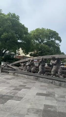 The video shows a series of stone sculptures of people carrying a large beam. The sculptures are arranged in a line, with each person in a different pose of exertion. The sculptures are in an outdoor setting, with trees and buildings visible in the background. The sky is cloudy. The pavement in front of the sculptures is made of large, square tiles. The camera pans across the sculptures from left to right, providing a view of each individual sculpture and the overall arrangement.
