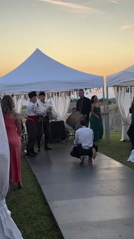 At an outdoor event with white tents against a sunset sky, a group of people are gathered. In the foreground, a man in a white shirt and dark pants crouches on a raised black platform, aiming a camera. Two men in traditional attire, one holding a staff, stand to his left on the platform. To the right of the crouching man, another man in a dark suit holds a large drum. Guests in dresses and suits are visible around the tents, and a woman in a green dress stands near a tent in the background.