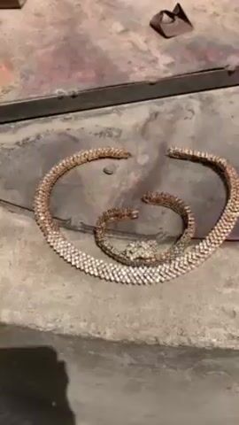 A close-up shot shows a gold-colored snake necklace and bracelet set on a flat, dirty surface that appears to be a car hood or other metal surface. The necklace is arranged in a semi-circular shape, with the bracelet placed inside the curve, mimicking the snake coiled. Both pieces are made of interlocking links embedded with small, shiny, clear stones, resembling diamonds or rhinestones. The head of the snake on both pieces is also detailed with the same stones, making it look like the snake is bejeweled. The surface beneath the jewelry is weathered and has patches of rust or dirt. The lighting suggests the scene is outdoors with natural sunlight.