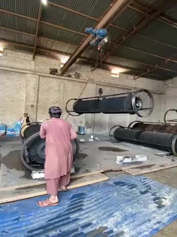 The scene is a large indoor warehouse or factory setting, characterized by its high corrugated metal roof and concrete walls. Multiple individuals are visible in the space, engaging in what appears to be an industrial process. A man dressed in a pink robe is actively involved, pushing a large, cylindrical black object that resembles an industrial drum. Above him, a motorized crane is suspending another similar drum in mid-air, presumably moving it into position or transferring it to another location. Other individuals are scattered throughout the area, some wearing masks, suggesting safety precautions are being taken. To the left, a pile of bagged material is visible, along with a large blue container. The floor is partially covered by a blue tarp, adding to the industrial setting. The overall impression is that the video captures an ongoing process within a manufacturing or industrial facility.