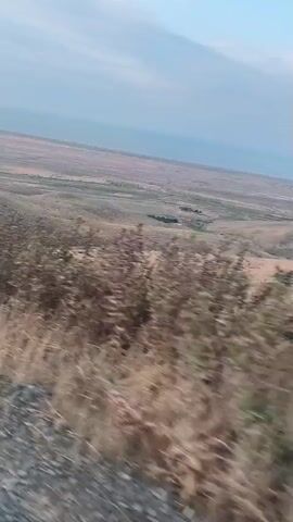 A first-person perspective video shows a landscape with dry grass, bushes, and hills under a hazy sky. The camera appears to be moving, possibly in a vehicle, as the foreground is blurred. The terrain is mostly brown and yellow, with some green vegetation scattered throughout. The horizon is indistinct, with the sky blending into the distant landscape. There are no people or discernible man-made objects visible. The overall impression is one of a dry, possibly arid environment.