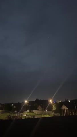 The video shows a nighttime aerial view of a residential area. Two streetlights illuminate the foreground, casting long beams of light across the scene. In the distance, the lights of houses and more streetlights are visible. The sky is dark and cloudy, with two bright points of light, possibly stars or planets, visible in the upper portion of the frame. The scene is static, with no movement or activity captured. The overall impression is one of a quiet, dimly lit evening in a rural or suburban setting.