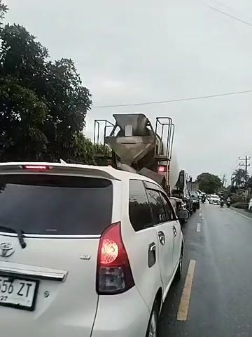 The video depicts a white car stopped in traffic behind a large, grey cement mixer truck. The truck is heavily soiled with cement residue. The camera slowly pans from the back of the car towards the rear of the cement truck. The sky is overcast. Other cars are visible further ahead in the traffic line. Trees line the side of the road. The road appears to be wet.