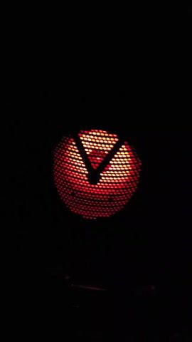 Here is a description of the video:

The video shows a circular object in a dark environment. The circle is made up of small, regularly spaced points that glow with light. Initially, the light is a warm yellow, but it changes to a bright blue and then goes dark. The change in color is sudden, alternating between the two colors. The shape within the circle appears to be an inverted V-shape. Below the circle, there are blurry metallic or reflective objects.