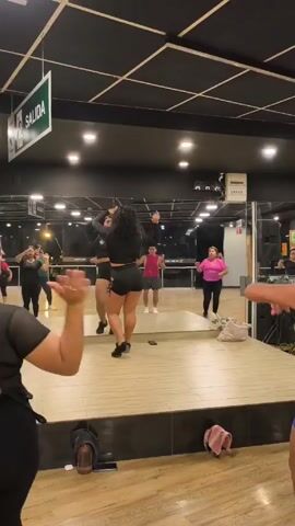 The video shows a group of people doing a dance workout in a mirrored studio. There are about ten people, including both men and women, in various athletic outfits. They are following a dance leader on a raised platform, with a DJ setting up equipment in the corner. The room has a black ceiling with fluorescent lighting and a wooden floor. The sign says "SALIDA" in green and white. There is a clock on the wall showing "GYM TIME" and the time is 20:03:11. There are some personal items like towels and bags scattered in the room.