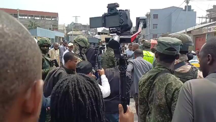 The video shows a crowded street scene. Many people are present, including individuals in military uniforms and construction workers with hard hats and yellow vests. There is a camera mounted on a gimbal being operated amidst the crowd. The surroundings appear to be an urban area with buildings of varying heights, some under construction. Overhead, there are power lines stretching between the buildings. The weather appears overcast.