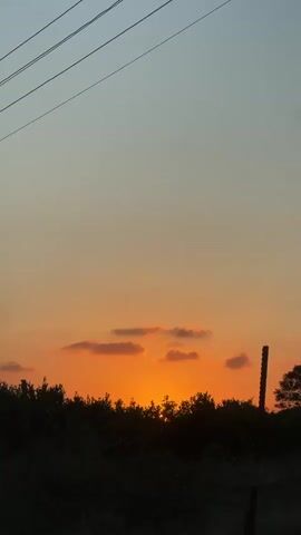 This video captures a sunset in Beit Hanan, Israel. The scene is dominated by the vibrant orange and yellow hues of the setting sun, which is partially obscured by a silhouetted line of trees and bushes. Scattered, light pink clouds drift across the sky, adding texture and depth to the sunset. Above the colorful sky, a lighter, pale blue gradient fades into a soft gray.

In the foreground, the dark silhouettes of vegetation create a stark contrast against the bright sky. A dark, vertical object, possibly a fence post or a utility pole, is visible on the right side of the frame. Overhead, thin, dark power lines stretch across the top of the image, indicating a populated or developed area.

The time of day is clearly sunset, as evidenced by the low angle of the sun and the rich, warm colors illuminating the sky. The weather appears clear, with only a few wispy clouds. The overall mood is tranquil and picturesque, evoking a sense of peace and natural beauty. There is no visible text in the media file.