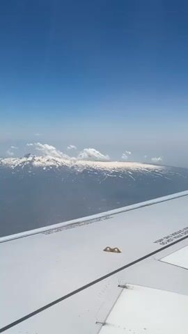 The video shows a view from an airplane window. The wing of the plane is in the foreground, and it is white with black lines. In the background, there is a mountain range covered in snow. The sky is blue with some white clouds. The camera is slowly panning to the right. There are no people visible in the video.
