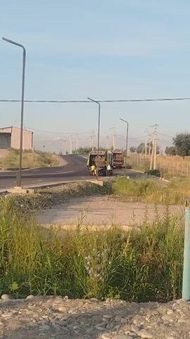 The video shows a rural road scene. In the foreground, tall green grass and some small purple flowers are visible. The middle ground features a dirt path leading to a paved road that curves to the left. Two dump trucks are on the paved road, with one truck in front of the other. Both trucks appear to be carrying loads in their beds. A person is visible between the two trucks, possibly operating or interacting with them. Two tall streetlights are positioned along the side of the road. In the background, there's a building with a gray roof and light-colored walls, along with utility poles and power lines, suggesting a somewhat developed but still rural environment. The sky is clear and blue, indicating daytime.
