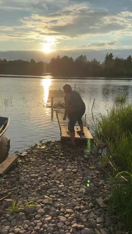 A person in a striped shirt and dark shorts is on a wooden dock by a body of water. The sun is setting in the background, casting a warm glow on the scene. The person is leaning over the edge of the dock, appearing to interact with the water. There is a small boat moored to the left of the dock. The shoreline is covered in pebbles and tall grass. The sky is cloudy, with the sun visible between the clouds.