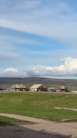 The video shows a rural landscape. There are several wooden houses lining a road, separated by colorful wooden fences. Behind the houses, a hill or mountain range stretches across the horizon. The sky is blue with fluffy white clouds.  The foreground features green grass and an asphalt road.  The camera pans slowly from left to right.