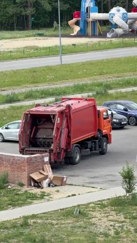 A red garbage truck with its rear lift mechanism open is parked in a lot near a brick structure overflowing with cardboard boxes. Several cars are parked nearby, including a white sedan, a dark blue sedan, a gray hatchback, and a white hatchback. In the background, a colorful children's playground with rocket-shaped structures is visible.
