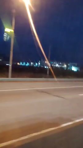 A nighttime video shows a view from a moving vehicle, likely a car or bus, traveling on a highway at dusk. The sky is a deep blue, transitioning to black, with a few faint lights from distant buildings visible on the horizon. The road is asphalt with white lane markings. A concrete barrier runs along the side of the road, separating it from the embankment. There are streetlights and utility poles visible. A billboard with an image of what appears to be nature is also seen. In the foreground, there are streaks of light and blur from the movement of the vehicle and the ambient lighting. A blue vehicle is partially visible in the right side of the frame, moving in the same direction.