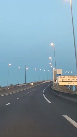 A first-person perspective video captures a view from a moving vehicle on a highway at dusk. The road ahead curves to the right, illuminated by streetlights that line the road on both sides. A single car is visible in the distance, traveling in the same direction. A large white sign with blue text and symbols is prominently displayed on the right side of the road. The sign indicates "AIRE DE LA BAIE DE SEINE" and shows symbols for parking and information, along with a distance marker of "2500m". Beyond the sign and the immediate highway infrastructure, a distant bridge can be seen. The sky is a pale, clear blue, transitioning from daylight to evening. The overall atmosphere is calm and serene.