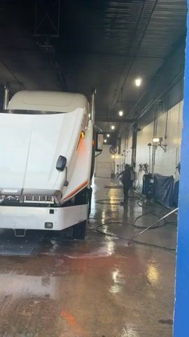 The video shows a white semi-truck with its hood open in what appears to be a commercial truck wash. A person wearing headphones and holding a phone stands near the truck, looking down at the device. In the background, another person is seen spraying the truck with a hose. The environment is an indoor bay with a concrete floor, overhead lighting, and industrial equipment visible along the walls. Water is pooling on the floor, indicating the washing process is underway.