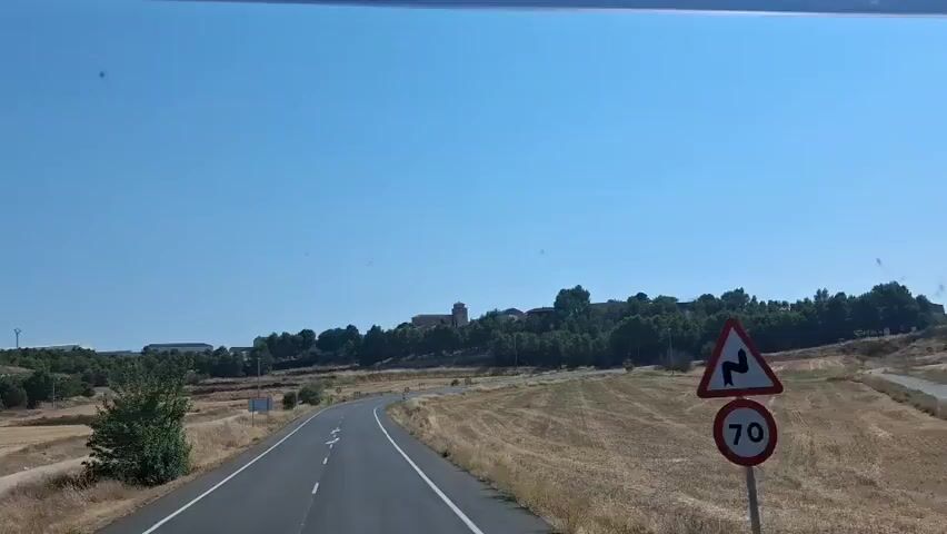 The video shows a view from a moving vehicle on a road with a clear blue sky above. The road curves to the left, with dry, yellowish grass and some trees on either side. In the distance, on a slightly elevated area covered with trees, there are buildings, including a prominent tower with a cross on top, suggesting a settlement or town. The fields on the right appear to have been harvested, with stubble visible. There are a few road signs visible, one with a circular red and white symbol. The overall environment is rural and dry, likely experiencing warm weather.