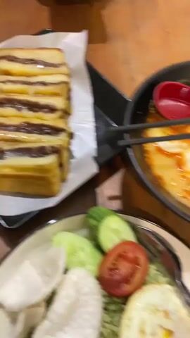 A close-up shot of two dishes of food on a wooden table. In the foreground is a plate with green rice, topped with a fried egg, cucumber slices, tomato slices, and white crackers. Behind it is a black bowl filled with ramen noodles and broth, garnished with a halved hard-boiled egg, seaweed, and what appears to be a slice of meat. A red spoon and chopsticks rest on top of the ramen. To the left, partially visible, is a stack of toasted bread with a dark filling, resting on white paper. In the background, out of focus, a person wearing a hijab is looking down at a smartphone. The lighting is warm and appears to be indoors.
