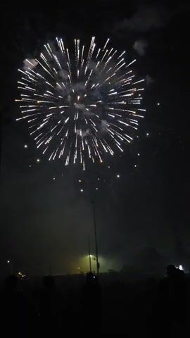 A fireworks display is visible against a dark night sky. The fireworks explode in various colors and patterns, including white, green, and red bursts that create glowing trails. In the foreground, silhouetted figures of people stand watching the spectacle. Several tall light poles are also visible, their lights illuminating the smoke from the fireworks. The overall atmosphere suggests a public event or celebration.