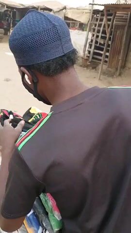 Here's a description of the video:

The video shows a person from the back, examining socks on a display table at an outdoor market. The person is wearing a dark blue kufi cap, a black face mask, and a black t-shirt with green, yellow, and red stripes on the shoulder. The socks on the table are in a variety of colors and patterns, many of them packaged in clear plastic. The market setting has other stalls and people visible in the background. A motorcycle and construction materials are also visible. The ground appears to be dirt.