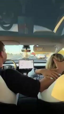Inside a Tesla car driving on a highway, a man is driving while looking at a woman sitting in the passenger seat. The man is wearing a black shirt, and the woman is wearing a light blue shirt. The man is singing along to music and laughing. Other cars are visible on the highway through the front windshield.