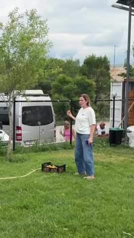 A woman with blonde hair pulled back is standing in a grassy area. She is wearing a white t-shirt and baggy blue jeans. She holds a yellow object, possibly a fruit, in her right hand near her mouth. In front of her on the grass is a black plastic crate filled with more yellow objects. To her left, a silver van is parked. Behind the van, a woman in a pink top is partially visible. To the right of the woman, a fence runs across the background. Behind the fence, there are some buildings and trees. The sky is overcast.