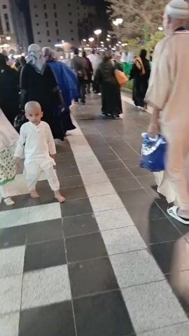The video shows a crowded outdoor area at night, possibly a public square or walkway, with people walking in various directions. Most of the people appear to be of Middle Eastern or South Asian descent. Several are wearing traditional Islamic clothing, including hijabs, abayas, and thobes. Two young children, a boy and a girl, are visible in the foreground. The boy is wearing a white thobe and is barefoot, while the girl is wearing a white top, a green patterned skirt, and a white head covering. They are walking hand-in-hand. The ground is paved with a pattern of light and dark gray tiles. In the background, there are buildings, streetlights, and trees. The overall atmosphere suggests a busy and possibly religious gathering.