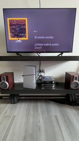 A static, eye-level shot depicts a home entertainment setup. A large, wall-mounted JVC television displays album art and lyrics from a Soda Stereo performance. Below, a dark gray shelf sits atop a barbell with weight plates on each end, creating a unique stand. The shelf holds a PlayStation 5 console, audio components including a turntable and amplifier, and two small, wood-cased speakers on either side. The floor is light gray laminate.