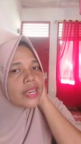 A medium shot shows a woman wearing a light pink hijab looking directly at the camera, with her left hand touching her jawline.  Behind her, there's a white wall, a red door with white blinds and a window with a red curtain.