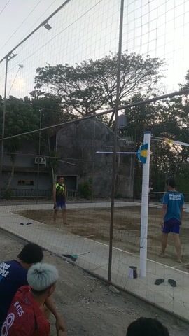 A volleyball game is underway on an outdoor court with a dirt surface. A net divides the court. Four individuals are visible on the court, with at least two more on the sidelines. The players are dressed in athletic attire, with one wearing a blue t-shirt and shorts, another a green and yellow tank top and shorts, and a third in a blue shirt and shorts. The sky is overcast, and trees are visible in the background, along with a building and some lighting fixtures. The camera is positioned low and angled upwards, and the video is filmed in portrait orientation.